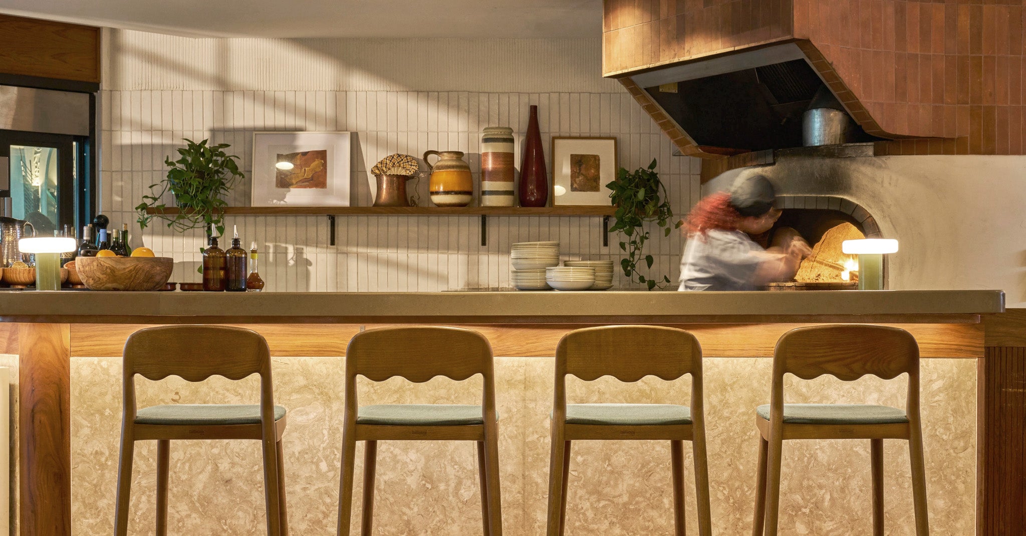 A warm, rustic kitchen interior with a wood-burning oven in the background, a stone countertop, and four wooden bar stools in the foreground.