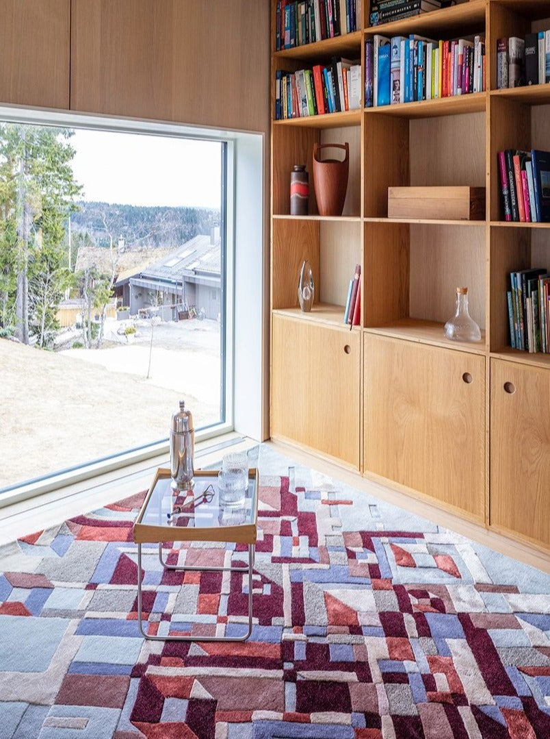 Stråler Red Brown  Modern interior with wooden bookshelf, colorful geometric rug, and view of nature outside.